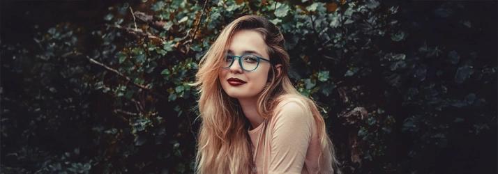 A girl in teal glasses. She sitting in front of a wall of greenery.